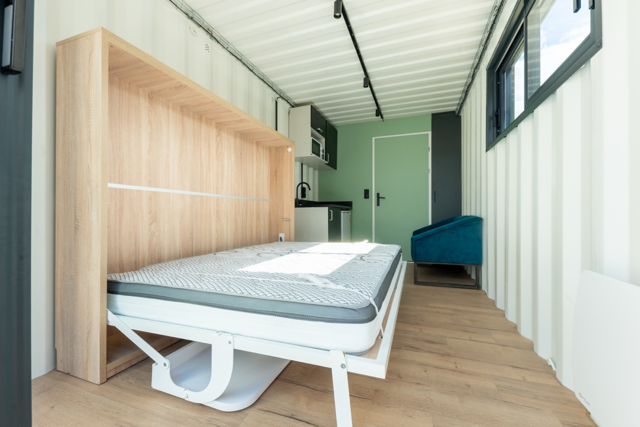 Intérieur EasyLogis avec lit escamotable ouvert, fauteuil et kitchenette au fond